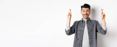 İyimser genç erkek, hayallerinin gerçekleşmesini umuyor, parmaklarını çapraz tutuyor, gülümsüyor ve iyi haberler bekliyor, beyaz arka planda duruyor..