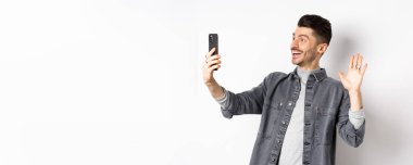 Genç adam cep telefonuyla görüntülü konuşma, akıllı telefon kamerasına el kaldırma, arkadaşınla internette selamlaşma, beyaz arka planda durma..