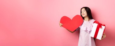 Sevgililer Günü konsepti. Aşk hediyeleri tutan romantik kız, büyük kırmızı kalp kartı ve hediye kutusu, rüya gibi bakışlar, aşık olmak, şefkatle gülümsemek, pembe arka plan.