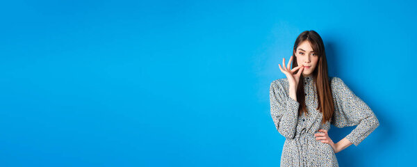 Serious woman promise to keep secret, seal lips, making zip gesture and looking at camera, standing in dress on blue background.