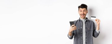 İnternetten alışveriş konsepti. İnternetten alışveriş yapan heyecanlı bir adam, plastik kredi kartı ve akıllı telefonla kolay satın alma, kameraya mutlu bir gülümseme, beyaz arka plan..