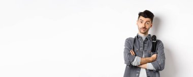 Ceketinin cebinde mikrofon tutan komik erkek şovmen ya da şarkıcı görüntüsü ve kollarını kavuşturup beyaz arka planda duran kameraya bakın..