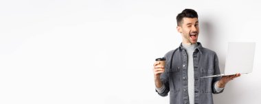 Kahve içen ve dizüstü bilgisayarla çalışan mutlu erkek işçi, kameraya göz kırpıyor, beyaz arka planda duruyor..