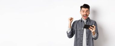 Heyecanlı adam cep telefonundan para kazanıyor ve seviniyor, elini kaldırıyor ve mutluluk ve neşe çığlıkları atıyor, beyaz arka planda akıllı telefonuyla duruyor..