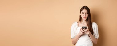 Akıllı telefonlu hayal kırıklığına uğramış, dudak büzen üzgün, telefonda kötü haberler okuyan, bej arka planda duran bir kız..