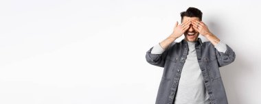 Yakışıklı mutlu adam gözlerini kapar ve şaşırmayı bekler, heyecanla gülümser, beyaz arka planda sıradan kıyafetler giyer..