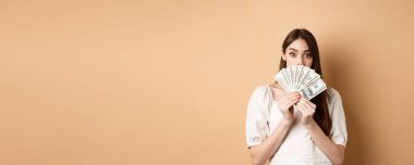 Dolarlar gösteren ve kaşlarını kaldıran heyecanlı kız hayret içinde, para kazanıyor, bej arka planda duruyor..