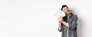 Mutlu zengin adam dolar banknotlarını kucaklıyor ve hayal kuruyor, parayla alışveriş yapmayı düşünüyor, beyaz arka planda duruyor..