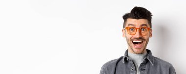 Heyecanlı, gözlüklü, gülümseyen komik adam, muhteşem tanıtıma hayran ve neşeyle bak, beyaz arka planda mutlu duruyor..