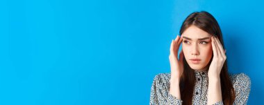 Genç bir kadının yakın plan portresi hasta hissediyor, baş tapınaklarına dokunuyor ve baş ağrısından çatık kaşlarını çatıyor, migren oluyor, logoya bakıyor, mavi arka plan..