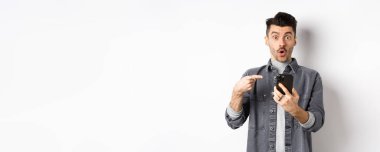 Vay canına, şuna bak. Heyecanlı yakışıklı adam telefonda teklif veriyor, akıllı telefonu işaret ediyor ve şaşkın bakıyor, beyaz arka planda duruyor..