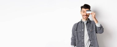 Bıyıklı mutlu yakışıklı adam gözlerinde plastik kredi kartı, kameraya heyecanlı bir şekilde gülümsüyor, beyaz arka planda duruyor..