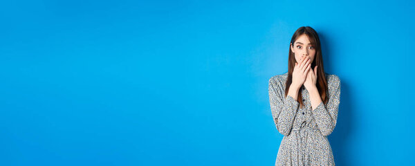 Shocked woman in dress gasping, covering mouth with hands and look excited, hear gossips, standing against blue background.
