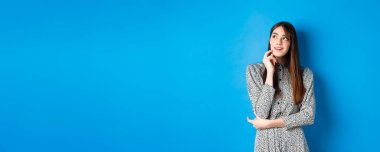 Yaratıcı genç bir kadın hayal ediyor ve gülümsüyor, sol üst logoya bakıyor düşünceli, bir fikri var, mavi arka planda duruyor..