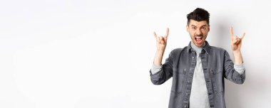 Salla gitsin. Küstah genç adam bağırıyor ve heavy metal boynuz işareti gösteriyor, harika bir konsere katılıyor, eğleniyor, beyaz arka planda duruyor..