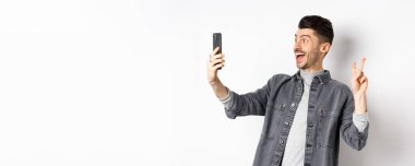 Akıllı telefondan selfie çeken neşeli bir gencin profili. Mobil uygulamayla fotoğraf çektirirken V işareti gösteriyor. Beyaz arka planda duruyor..