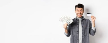 Elinde plastik kredi kartı ve dolar banknotları olan heyecanlı adam beyaz arka planda eğleniyor..