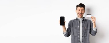 Plastik kredi kartı ve boş akıllı telefon ekranı, reklam uygulaması, beyaz arka planda duran heyecanlı yakışıklı adam..