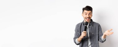 Mikrofonla konuşan, el kol hareketi yapan, mikrofonla konuşma yapan, beyaz arka planda duran yakışıklı bir sanatçı..