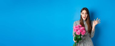 Sevgililer Günü konsepti. Aşığından çiçek buketi alan güzel bir kız kameraya şaşkın ve mutlu bakıyor, mavi arka planda duruyor..