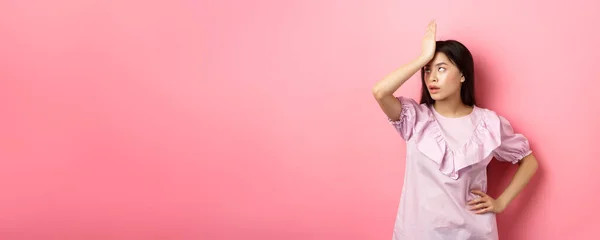 Annoyed asian girl roll eyes and facepalm, feeling tired and irritated with stupid conversation, standing against pink background.