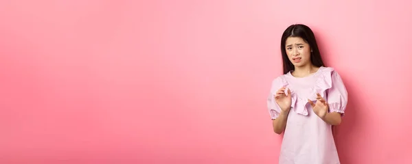 Stay away from me. Disgusted asian girl raising hands to block someone, cringe from aversion, look reluctant and asking to stop, rejecting something bad, pink background.