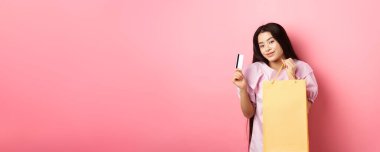 Alışverişe. Pembe arka planda duran, alışveriş çantası ve plastik kredi kartı gösteren şık Asyalı kız..