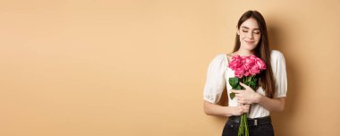 Sevgililer Günü. Şefkatli ve romantik Kafkasyalı kadın, gözleri kapalı, rüya gibi, gizli bir hayran ya da sevgiliden bir buket gül, bej arka planda duruyor..