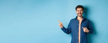 Mutlu gülümseyen adam sol logoyu işaret ediyor, reklamını yapıyor, mavi arka planda duruyor..