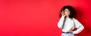 Image of cute girl with curly hair making promise to stay quiet, zipping lips, making seal, hiding a secret, standing dumb on red background.