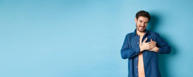 Gülümseyen adam duygulanmış ve minnet duyuyor, kalple el ele tutuşuyor ve teşekkür ediyor, mavi arka planda duruyor. Boşluğu kopyala