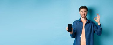 Yakışıklı genç adam iyi bir hareket ve boş akıllı telefon ekranı gösteriyor. Uygulama ya da şirket öneriyor. Mavi arka planda duruyor..