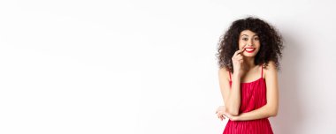 Kırmızı elbiseli, ısıran parmağı olan ve kameraya tatlı tatlı gülümseyen güzel bir kadın beyaz arka planda duran bir şey istiyor..