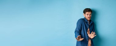 Çekingen genç adam uzak durmak istiyor, ellerini savunmaya uzatıp geri çekiliyor, iğrenç bir şeyi reddediyor, mavi arka planda duruyor..