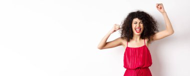 Neşeli, duygusal, kıvırcık saçlı, kırmızı elbiseli, elini kaldıran ve şarkı söyleyen, takımı destekleyen, kazanmak isteyen, beyaz arka planda duran bir kadın..