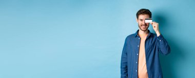 Mutlu erkek müşteri elinde plastik bir kredi kartı tutuyor ve gülümsüyor, mavi arka planda günlük kıyafetler içinde duruyor..