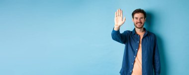 Dost canlısı genç adam gülümsüyor ve beşlik çakıyor, selam vermek için feragat ediyor, mavi arka planda duruyor..
