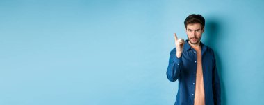 Ciddi bir genç adam azarlıyor, onaylamayan bir şekilde parmağını sallıyor, kötü bir şeyi uyarıyor veya yasaklıyor, mavi arka planda duruyor..