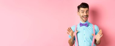 Papyonlu neşeli adam, pantolon askılarını gösteriyor ve gülümsüyor. Mutlu, neşeli, pembe arka planda heyecanlı duruyor..