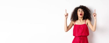 Gece makyajlı, kıvırcık saçlı, heyecanlı ve dik duran, reklam yapan, beyaz arka planda kırmızı elbiseli harika bir kız..