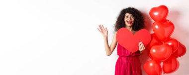 Sevgililer Günü ve aşk konsepti. Zarif kırmızı elbiseli neşeli genç bir kadın, romantik balonların yanında duruyor ve büyük kırmızı bir kalp kesiği tutuyor, merhaba demek için el sallıyor, randevuyu bekliyor..