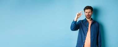 Kafası karışmış bir adam, küçük boyda, şüpheli ya da şüpheci görünüyor, mavi arka planda duruyor. Boşluğu kopyala