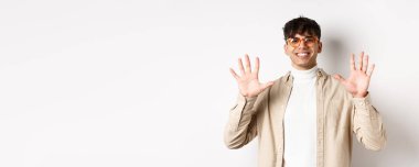Gözlüklü ve şık giyinmiş modern bir adam, on parmak numarası gösteriyor ve gülümsüyor, beyaz arka planda duruyor..