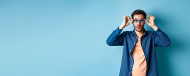 Komik genç adam yeni güneş gözlüğü deniyor, kameraya şaşırmış, mavi arka planda duruyor. Boşluğu kopyala