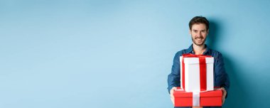 Sevgililer günün kutlu olsun. Yakışıklı bir adam kız arkadaşına hediyeler veriyor, hediye kutuları tutuyor ve gülümsüyor, mavi arka planda dikiliyor..