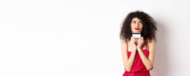 Romantik görünen genç bir kadın, elinde plastik kredi kartıyla alışverişi düşünüyor, beyaz arka planda dikiliyor. Boşluğu kopyala