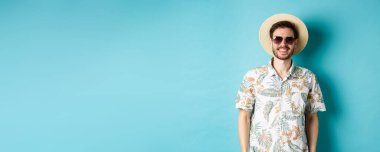 Güneş gözlüklü ve yaz gömlekli yakışıklı adam, turneden zevk alıyor, mavi arka planda duruyor..