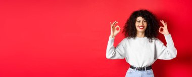 Kıvırcık saçlı, kendine güvenen güzel bir kız. Güzel jestler yapıyor, gülümsüyor, onaylıyor ve sana katılıyor. Mükemmel seçimi övüyor, kırmızı arka planda tatmin oluyor..