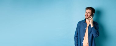 Modern gülümseyen adam cep telefonuyla konuşuyor, kameraya mutlu görünüyor, mavi arka planda duruyor..