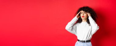 Sıkıntılı kıvırcık saçlı İtalyan kız, baş şakaklarına dokunuyor ve kötü hissediyor, baş ağrısı çekiyor, yana bakıyor, kırmızı arka planda duruyor..